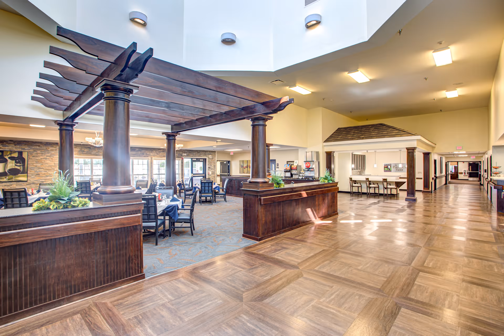 Spacious dining area in a senior living facility with wooden columns and beams forming a pergola-like structure over tables set for meals. The room has large windows allowing natural light, a high ceiling, and a long hallway leading to other parts of the facility.
