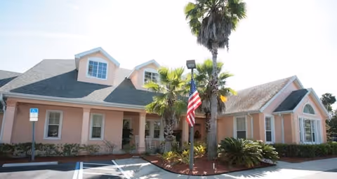 Front exterior of a peach-colored single-story senior living building with palm trees, an American flag, and a small parking area.