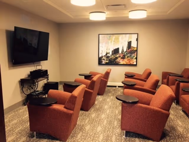 A small media room with multiple red upholstered armchairs arranged in rows facing a wall-mounted flat screen TV. Each chair has a small attached black table. The room has beige walls, a patterned carpet, ceiling lights, and a framed abstract painting on the wall behind the chairs.