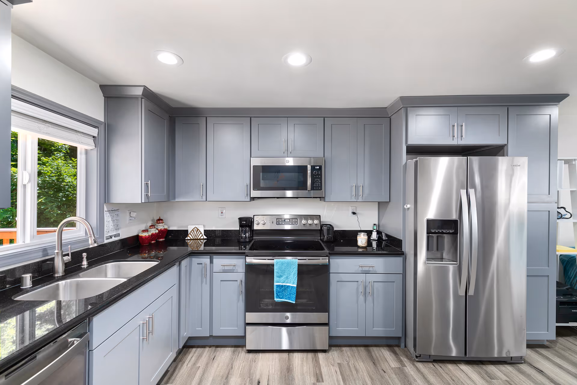 Modern kitchen with gray cabinets, black countertops, stainless steel appliances including a refrigerator, oven, and microwave. There is a double sink under a window with a view of greenery outside. The floor is light wood, and the kitchen is well-lit with recessed ceiling lights.