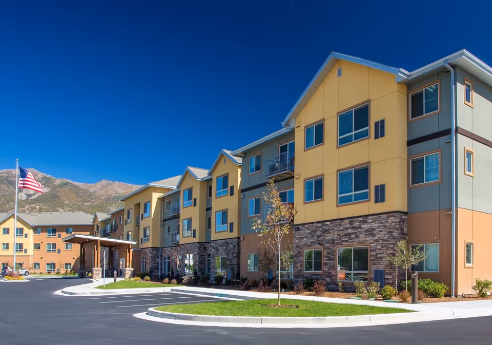 Exterior view of a modern three-story senior living facility building with yellow, gray, and brown walls, multiple windows, and a covered entrance. There is a small landscaped area with young trees and shrubs, a paved driveway, and an American flag on a flagpole. Mountains are visible in the background under a clear blue sky.