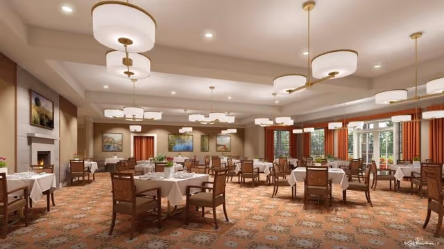 A spacious dining room with multiple round tables covered with white tablecloths and set with plates and glasses. The room features warm lighting from modern ceiling fixtures, large windows with orange curtains, framed artwork on the walls, and a fireplace on the left side. The carpet has a patterned design in warm tones.