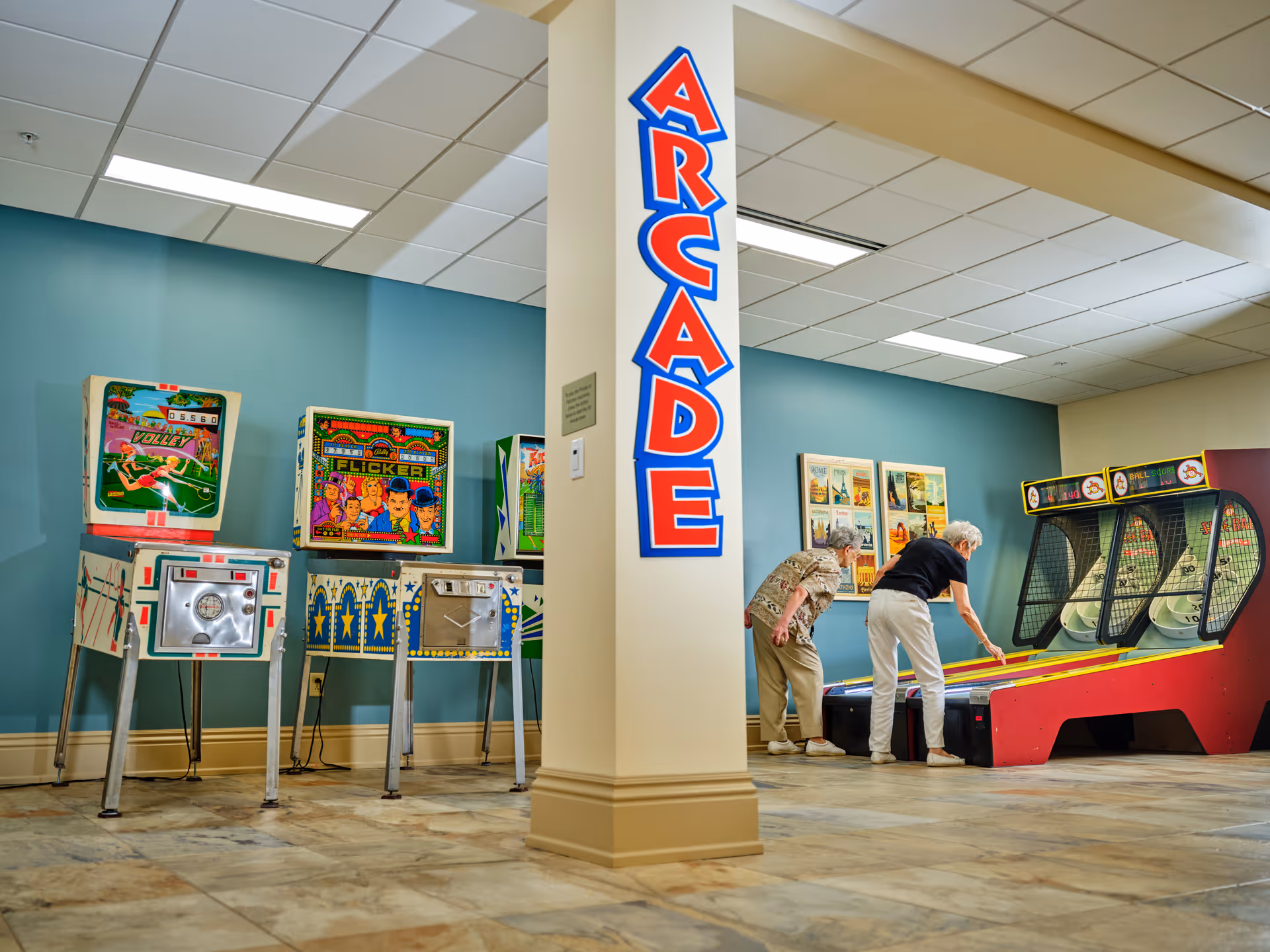 Two elderly women play skee-ball in an indoor arcade area with vintage arcade machines and a large sign reading ARCADE.