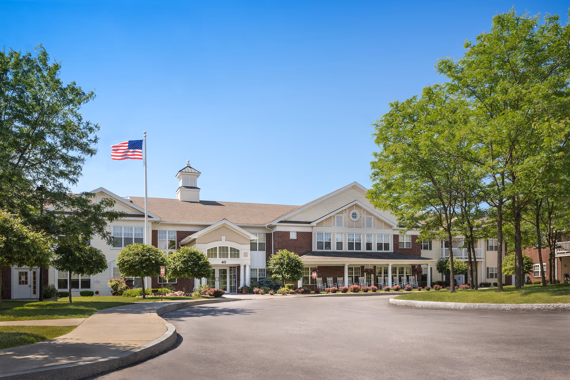 Front exterior view of The Addison of Willow Pond facility with a clear blue sky, an American flag on a flagpole, well-maintained landscaping including trees and bushes, and a curved driveway leading to the entrance.