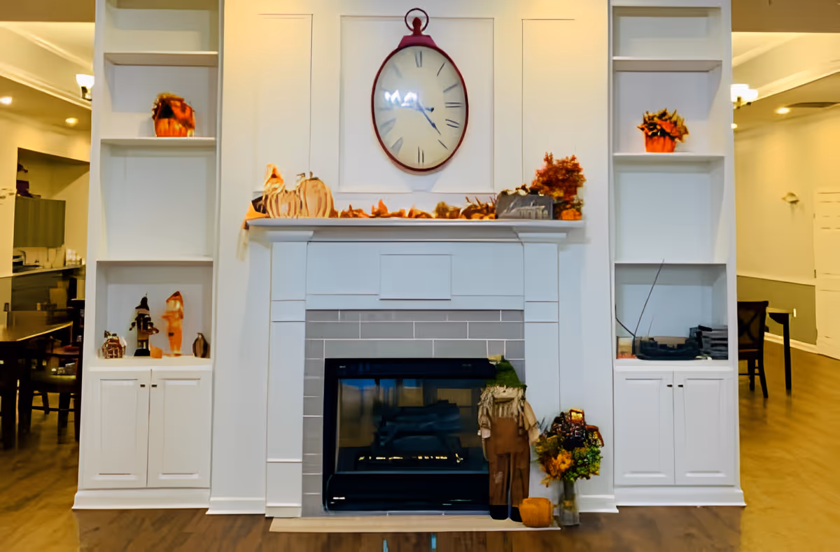 A cozy indoor fireplace with a white mantel and built-in shelves on either side. The mantel is decorated with autumn-themed items including pumpkins, leaves, and small plants. Above the mantel hangs a large round clock with Roman numerals. The shelves hold additional decorative items. The room has warm lighting and wooden flooring.
