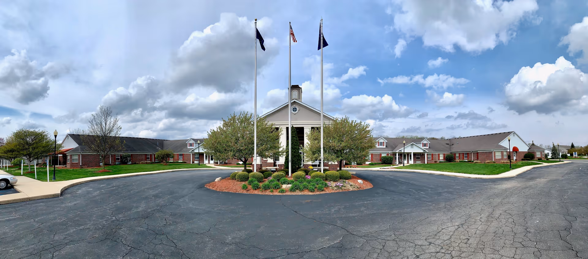 Wide exterior view of a single-story senior living facility with a circular driveway and three flagpoles in the center. The building has a brick facade with white trim and a pitched roof. There are trees and landscaped bushes around the entrance area under a partly cloudy sky.