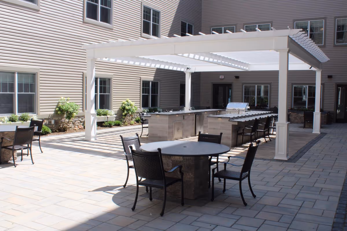 Outdoor courtyard area with a white pergola covering a built-in grill and bar seating. There are round tables with chairs arranged on a tiled patio, surrounded by the beige exterior walls and windows of the building.