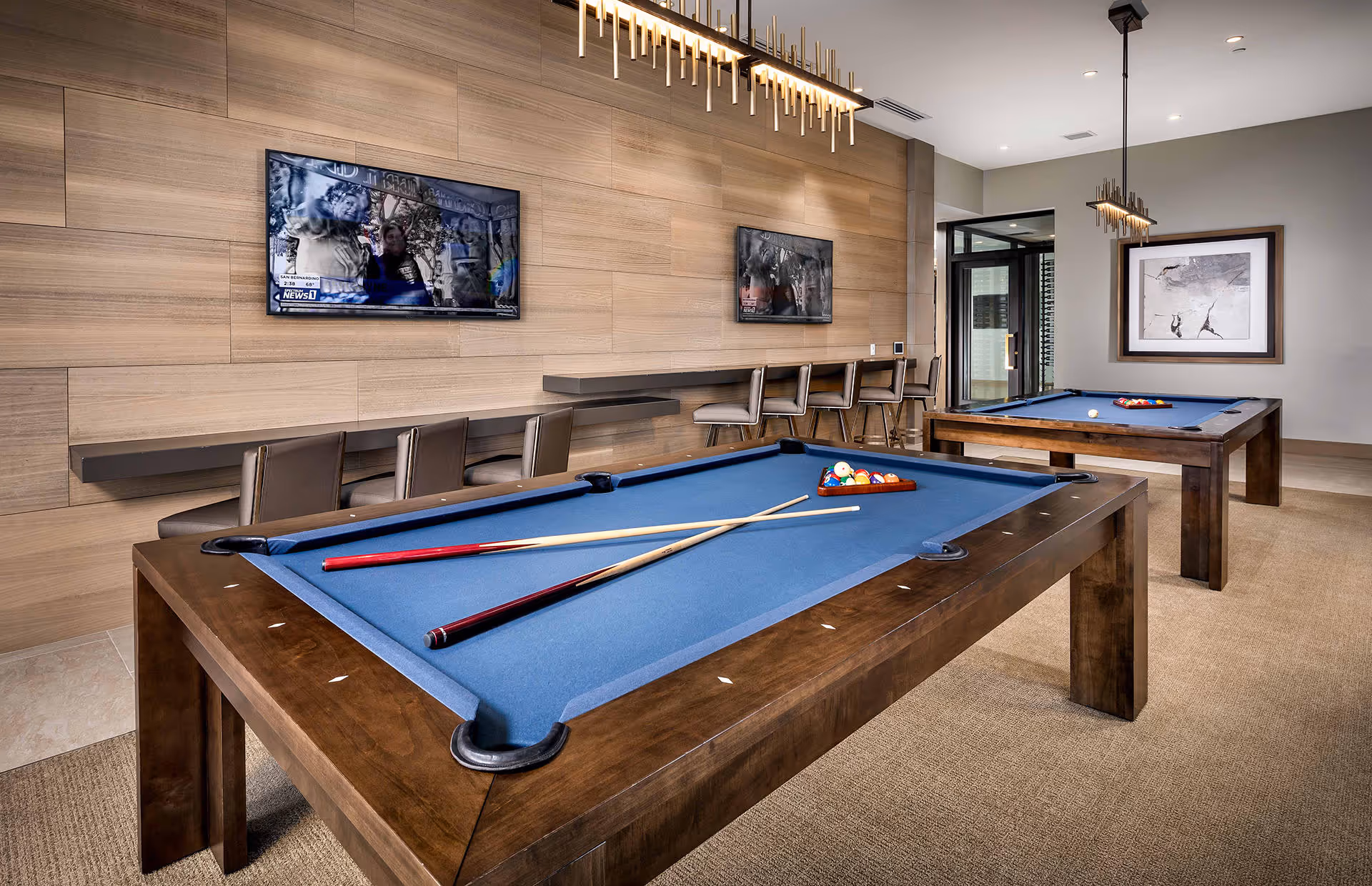 Interior of a recreational room featuring two pool tables with blue felt, pool cues, and balls arranged on the tables. The room has a modern design with wood-paneled walls, a row of bar stools along a counter, two wall-mounted flat-screen TVs, and contemporary pendant lighting. A framed abstract artwork is visible on one wall.