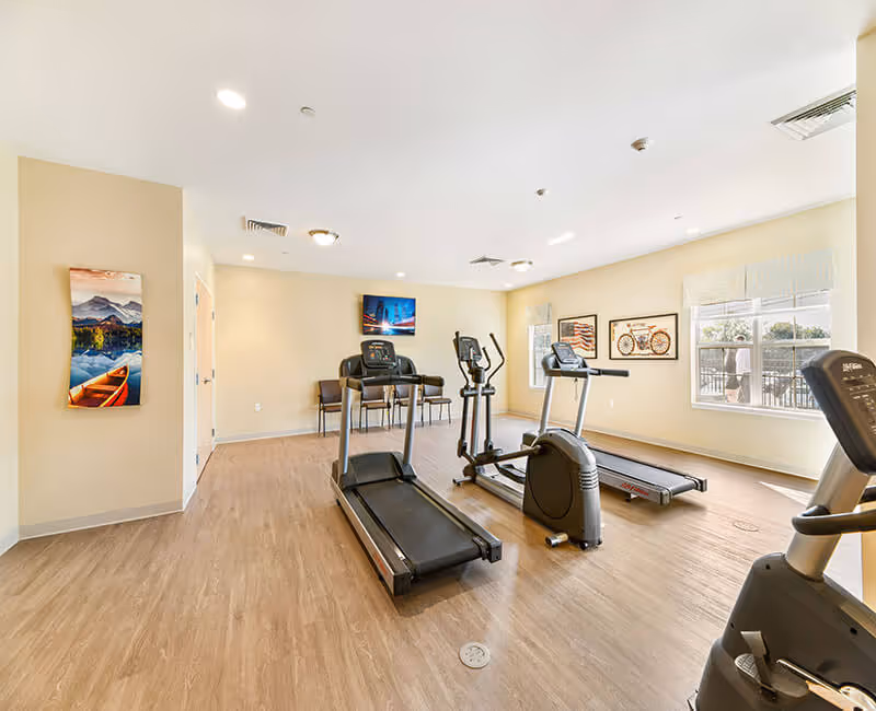 Bright fitness room with treadmills, an elliptical, an exercise bike, chairs, and wall art.