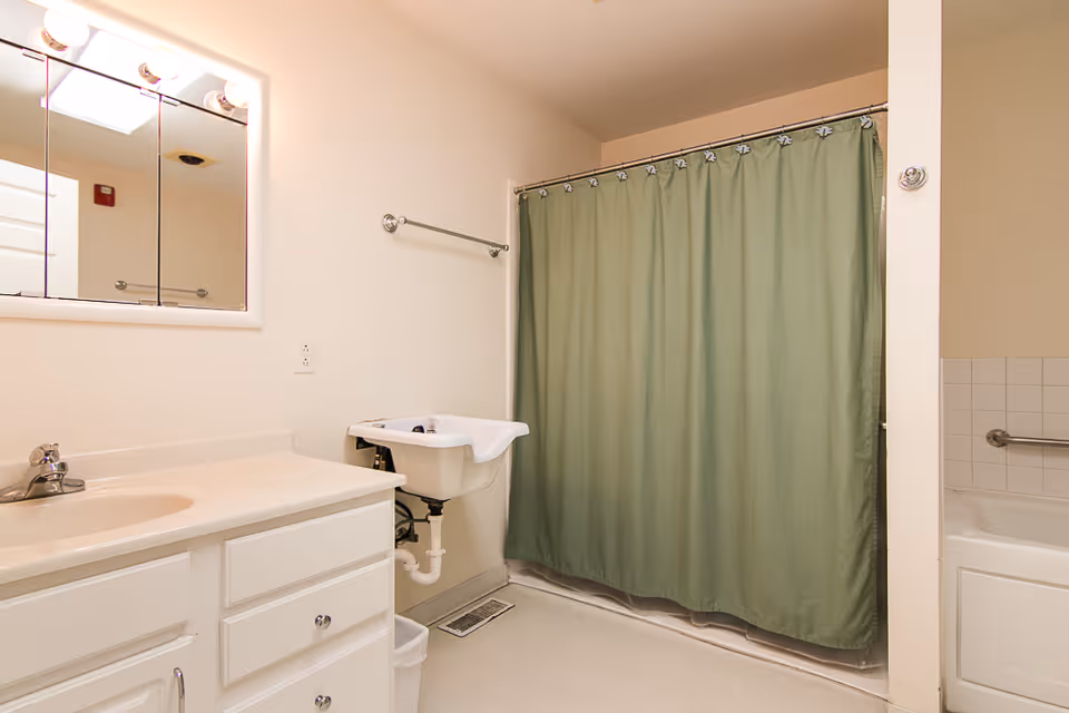 A clean bathroom with a white vanity and sink, a wall-mounted mirror with lights above it, a small white utility sink, a green shower curtain covering a bathtub, and beige walls.