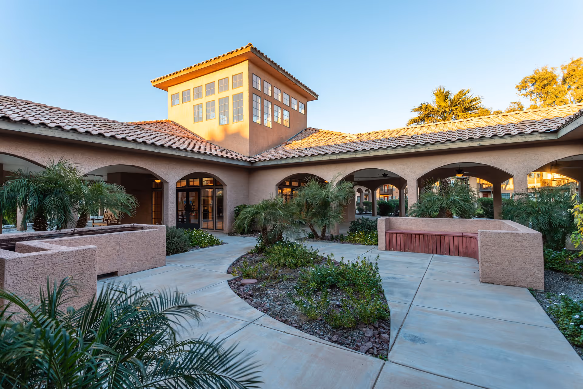 Outdoor courtyard area of a senior living facility with paved walkways, built-in seating, palm trees, and a building with arched doorways and windows under a tiled roof during daylight.