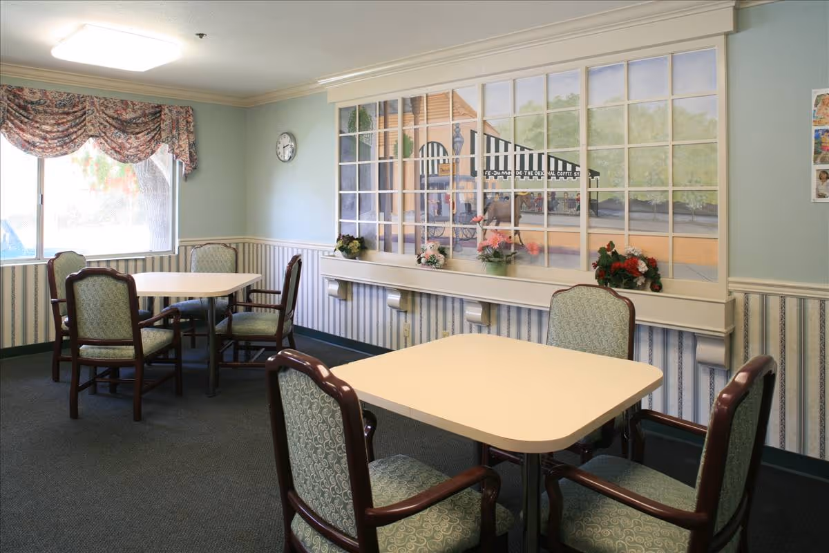 Small dining room with two square tables surrounded by upholstered chairs, a large painted mural 'window' on the wall, and floral decorations.