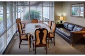 A cozy interior room with a rectangular table covered by a white tablecloth surrounded by six wooden chairs with cushioned seats. To the right, there is a dark brown leather sofa with a lamp on a side table next to it. Large windows with blinds cover the walls on two sides, letting in natural light and showing greenery outside.