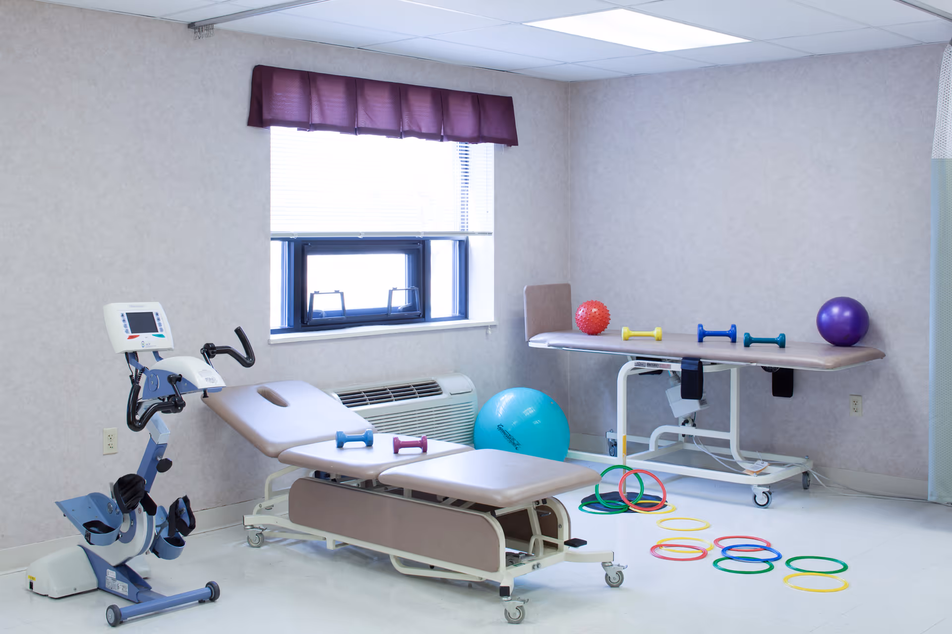 A physical therapy room with two padded treatment tables, an exercise bike, various colorful exercise balls, small dumbbells, and plastic rings on the floor. The room has a window with a purple valance and light-colored walls.