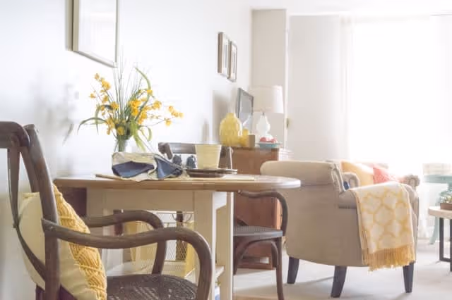 A bright and cozy living room area with a wooden dining table set with dishes and napkins, surrounded by chairs. There is a beige armchair with a yellow patterned throw blanket draped over it, a wooden sideboard with decorative items including a lamp and a vase with yellow flowers, and large windows letting in natural light.