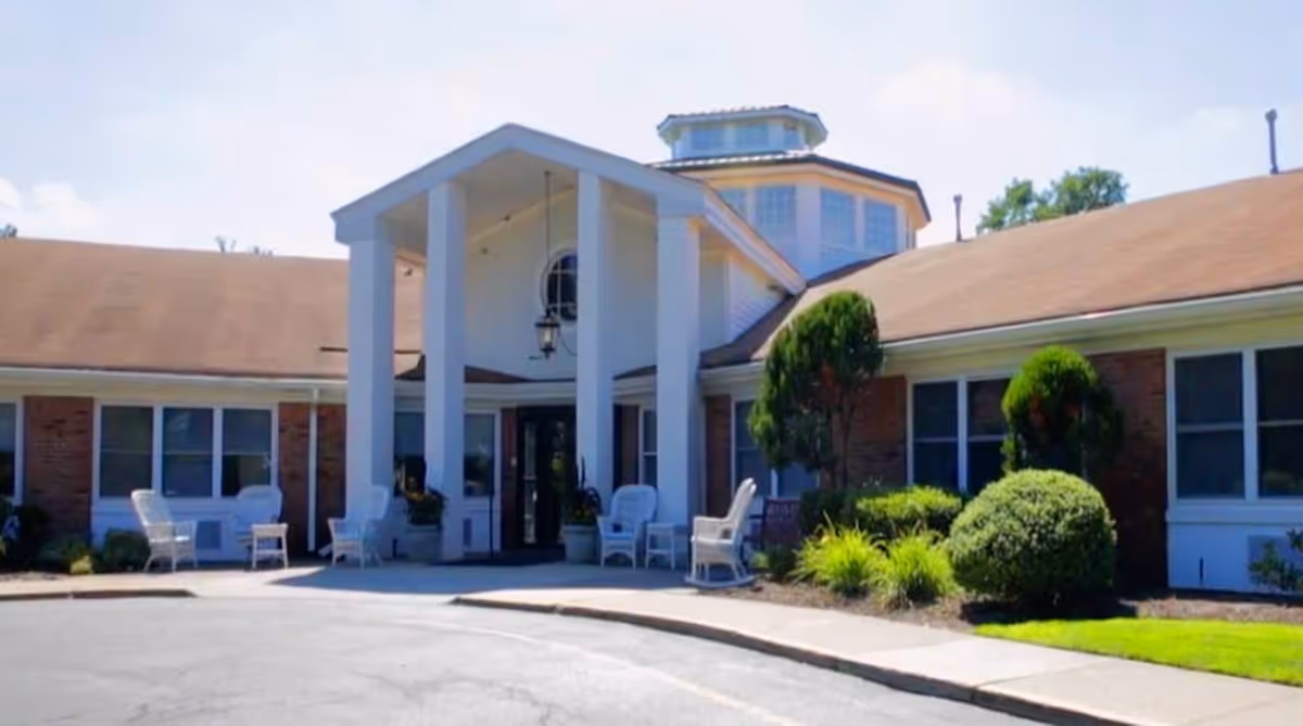 Front exterior view of CareOne at The Cupola, showing a single-story building with a covered entrance supported by white columns, surrounded by greenery and outdoor seating.