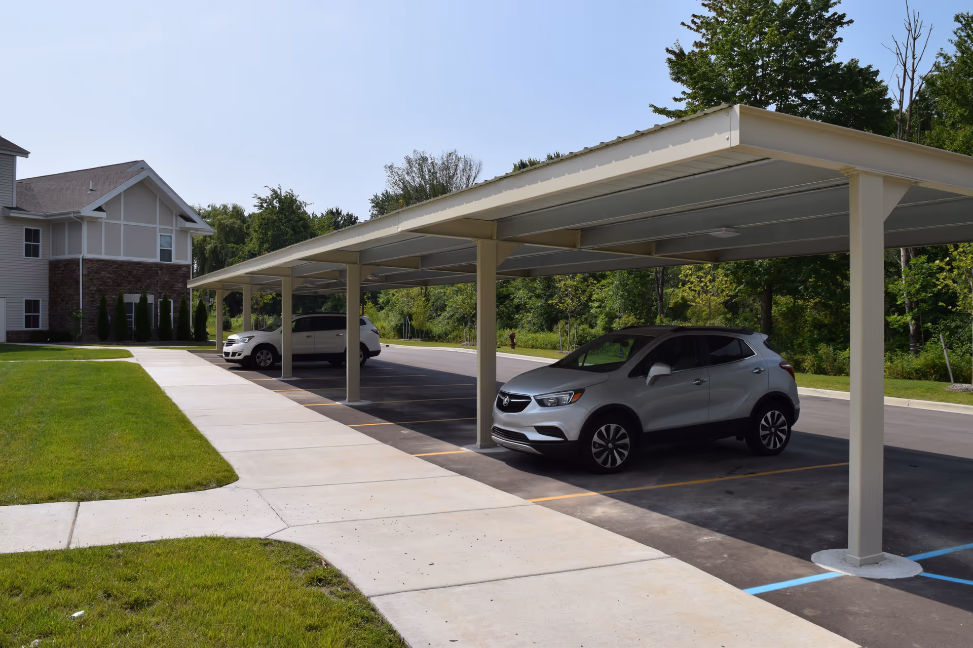 Covered parking area with two cars parked under a metal canopy next to a sidewalk and a building with stone and siding exterior, surrounded by green grass and trees under a clear sky.