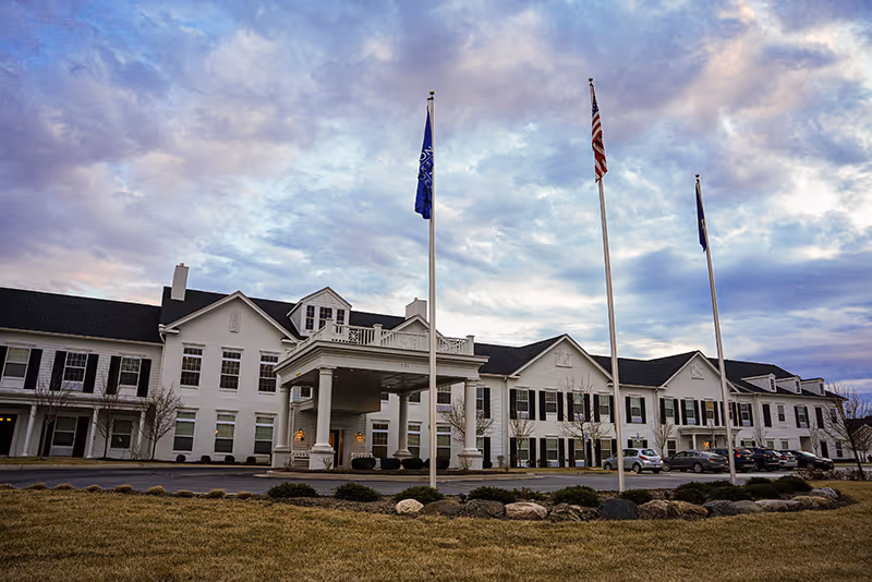 Exterior view of a large two-story senior living facility building with white siding and black shutters under a partly cloudy sky at dusk. Three flagpoles with flags stand in front of the building, and several cars are parked near the entrance.