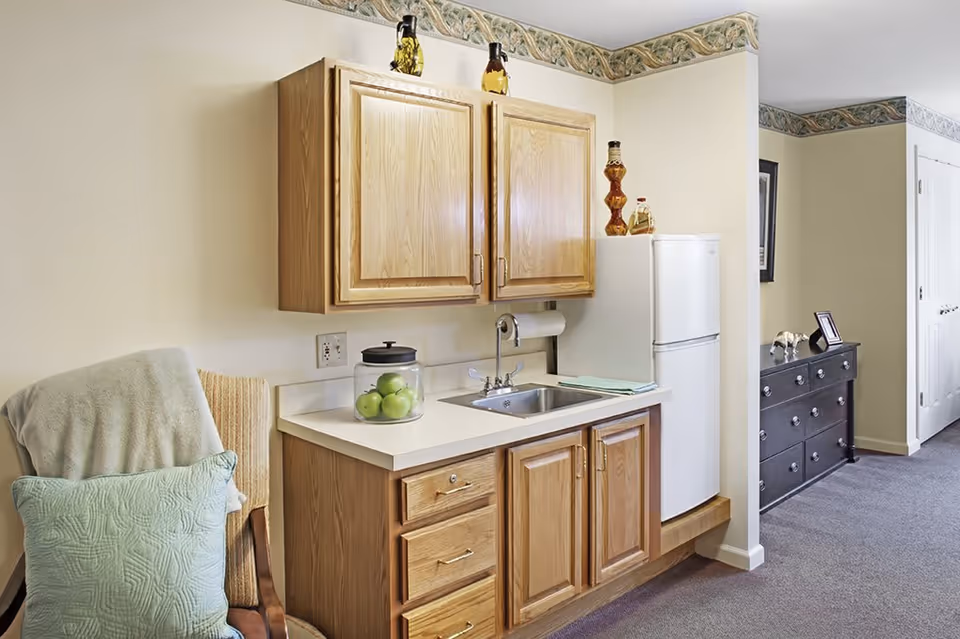 Small kitchenette area with wooden cabinets, a white countertop with a sink, a glass jar filled with green apples, and a white refrigerator. To the left, there is a chair with a green pillow and a beige blanket draped over it. The room has beige walls with a decorative border near the ceiling and a carpeted floor. In the background, there is a black dresser with decorative items and a framed picture on the wall.
