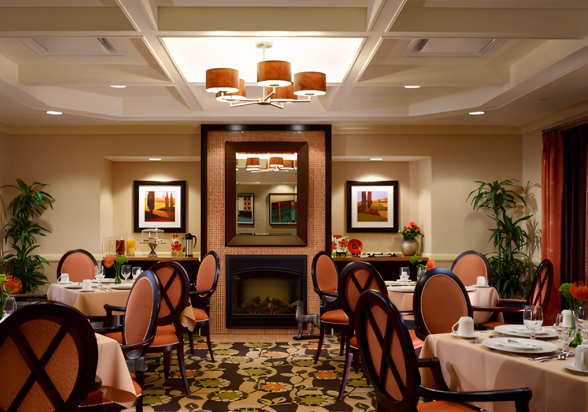 A warmly lit dining room with round tables covered in beige tablecloths, set with white plates, cups, and glassware. The room features cushioned chairs with wooden frames and orange upholstery. A fireplace with a large mirror above it is centered on the far wall, flanked by two framed paintings and potted plants. A modern chandelier hangs from the ceiling, and the carpet has a floral pattern.