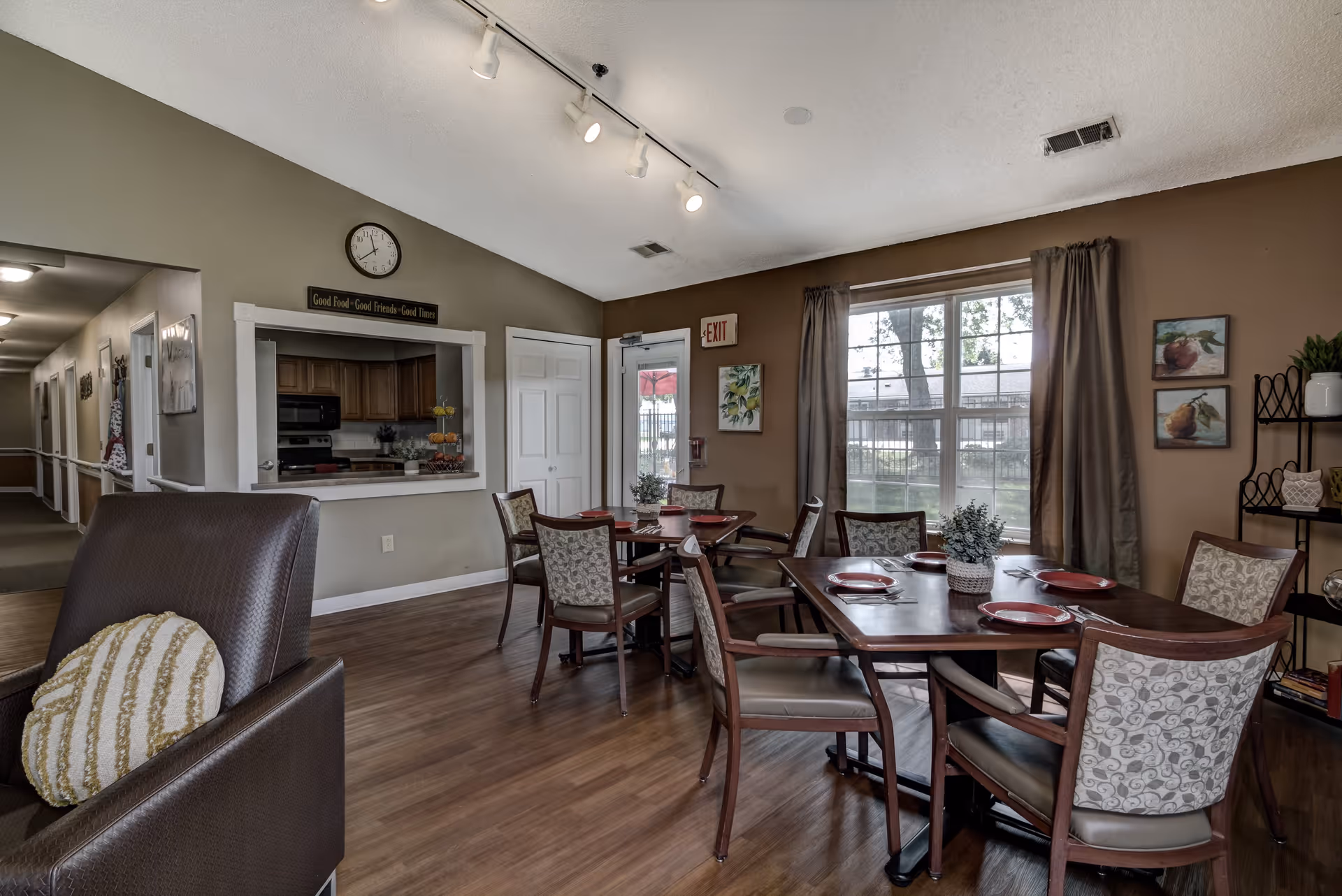 Dining area in a senior living facility with two wooden tables set with red plates and silverware. There are six upholstered chairs around the tables. A large window with brown curtains lets in natural light. To the left, there is a kitchen pass-through window with wooden cabinets and a clock above it. The walls are painted in neutral tones, and there is a hallway visible in the background. A brown armchair with a round cushion is partially visible in the foreground.