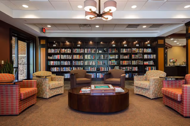 A cozy library or reading room with a large bookshelf filled with books along the back wall. In front of the bookshelf, there are six upholstered armchairs arranged in a semi-circle around a round wooden coffee table with books and magazines on it. The room has warm lighting with a modern ceiling light fixture and large windows on the left side letting in natural light.