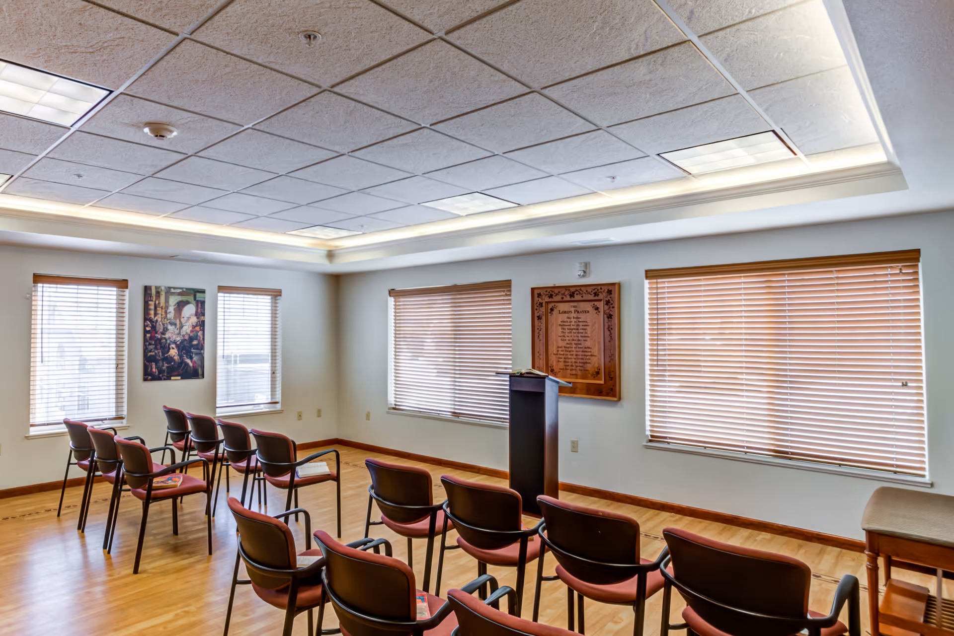 A small meeting or chapel room with rows of chairs facing a wooden podium. The room has wooden floors, several windows with blinds, a framed plaque on the wall, and a painting between two windows.