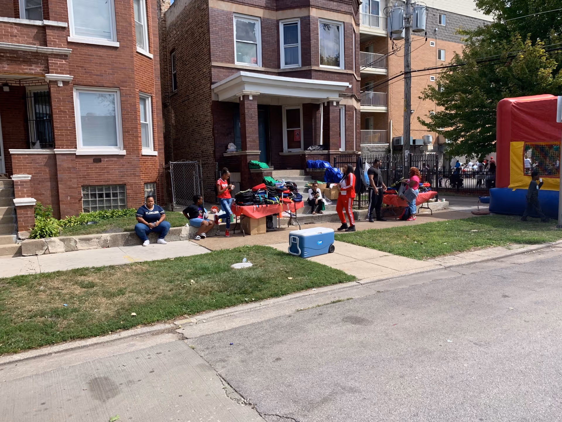 People gathered outside a brick residential building with tables set up on the sidewalk holding various items. Some individuals are sitting on the curb, while others stand near the tables. There is a blue cooler on the grass and a colorful inflatable bounce house to the right. Trees and other buildings are visible in the background.