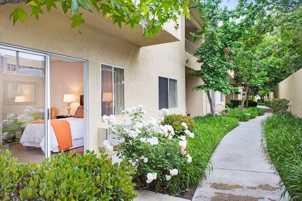 Outdoor walkway alongside a beige building with green bushes, white flowers, and trees. A bedroom with a bed, orange blanket, and lamp is visible through a sliding glass door.