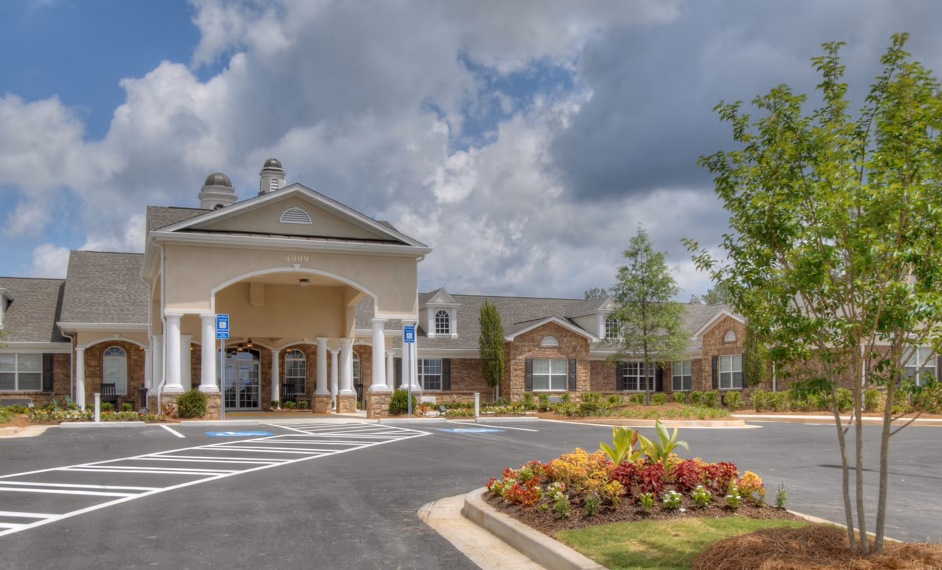 Front entrance of a single-story senior living facility with a covered portico, columns, parking lot, and landscaped flowerbeds under a cloudy sky.
