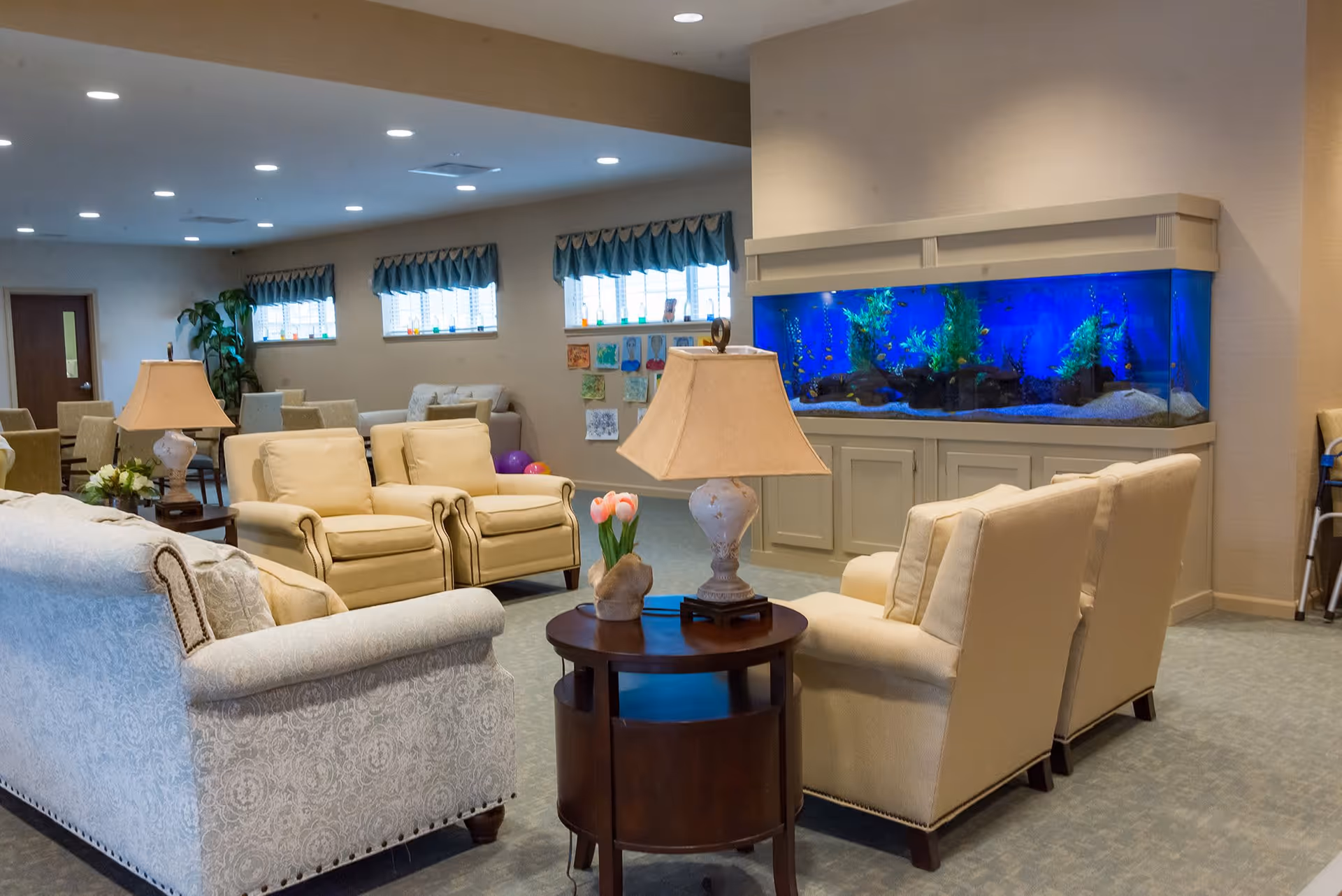 A cozy living room area in a senior living facility with beige armchairs and a patterned sofa arranged around a wooden side table with a lamp and a small flower vase. A large built-in aquarium with blue lighting is set into the wall, and there are small windows with valances along the back wall. The room has soft lighting and a carpeted floor.