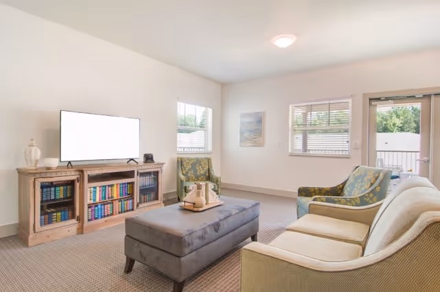 A bright living room with a beige sofa, two patterned armchairs, a gray upholstered ottoman in the center, and a wooden TV stand with books and decorative items. The room has two windows and a glass door leading outside, with light-colored walls and carpeted floor.