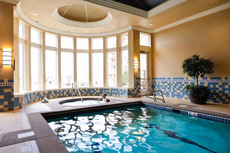 Indoor swimming pool area with a hot tub where a person is relaxing. The space has large windows allowing natural light to fill the room, tiled walls with blue and beige patterns, and a potted plant near the pool.