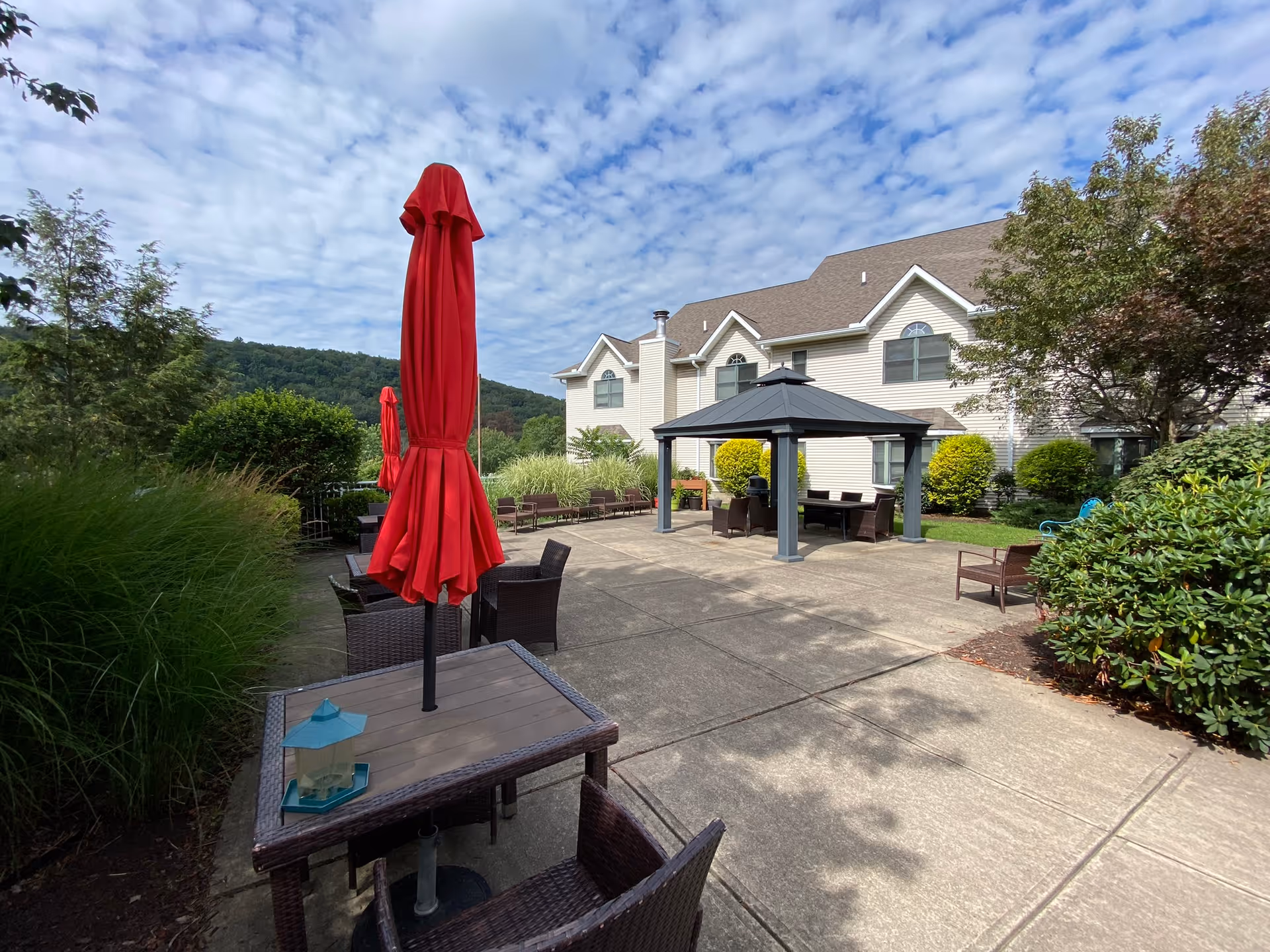 Outdoor patio area at Willowbrook Place with several wicker tables and chairs, some tables have closed red umbrellas. There is a gazebo with seating underneath, surrounded by bushes and trees, with a building and hills in the background under a partly cloudy sky.