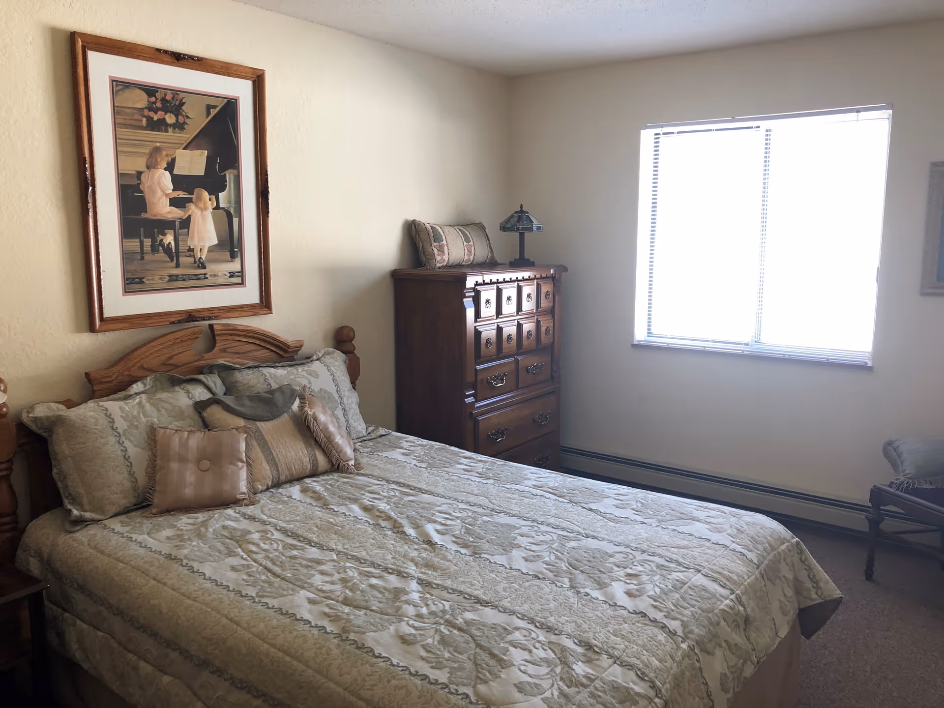 A bedroom with a bed covered in a patterned beige and green bedspread and several decorative pillows. Above the bed is a framed picture of two children playing a piano. To the right of the bed is a wooden dresser with a small lamp and a cushion on top. A window with blinds is on the far wall, letting in bright light. There is also a wooden chair partially visible on the right side of the image.