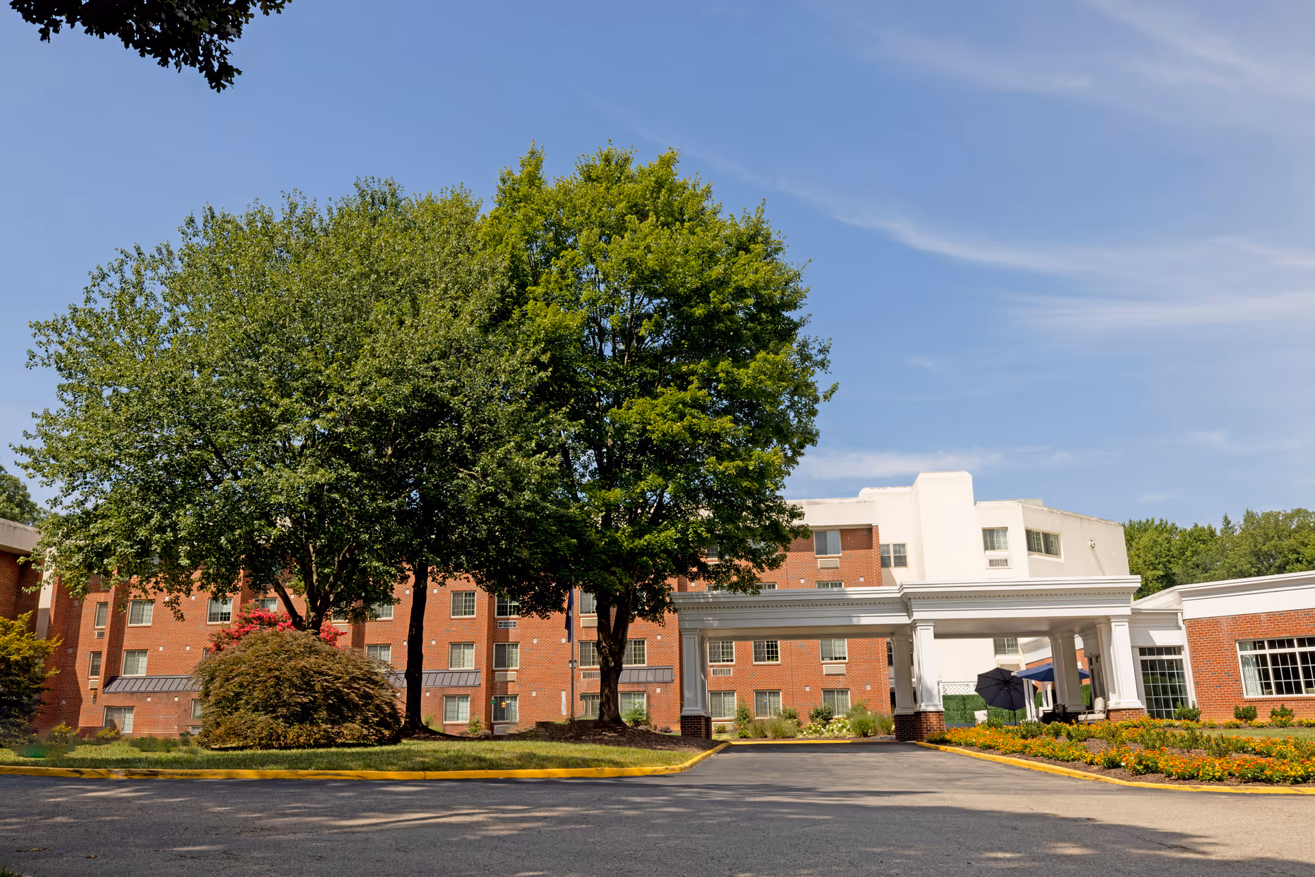 Exterior view of The Barrington at Hioaks senior living facility showing a multi-story brick building with white accents, a covered entrance supported by columns, landscaped flower beds, and large green trees under a clear blue sky.