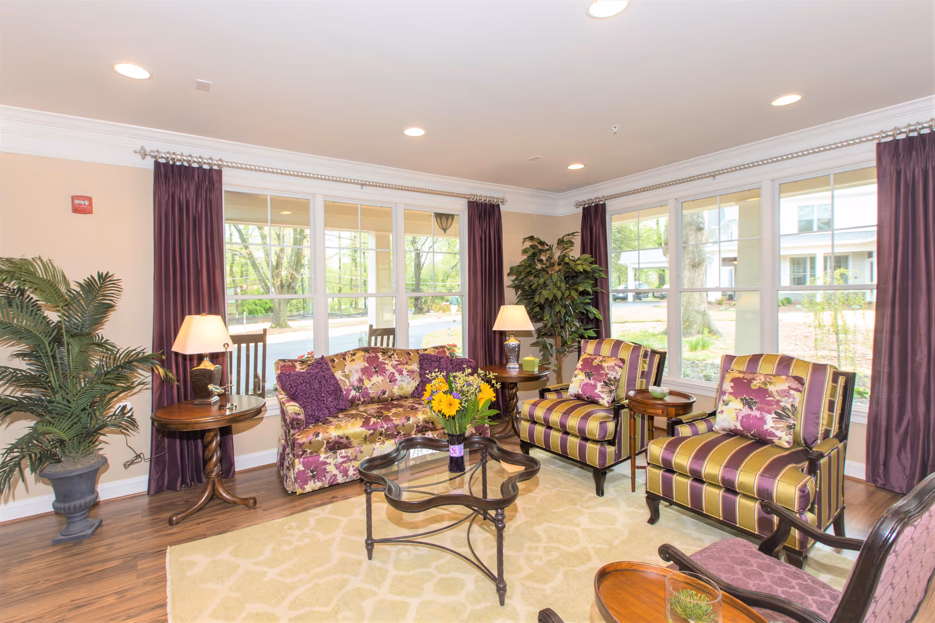 A bright and cozy living room with large windows letting in natural light. The room features a floral patterned sofa, two striped armchairs with floral cushions, a glass coffee table with a vase of yellow flowers, two side tables with lamps, and potted plants. The floor is wooden with a light-colored area rug, and purple curtains frame the windows.