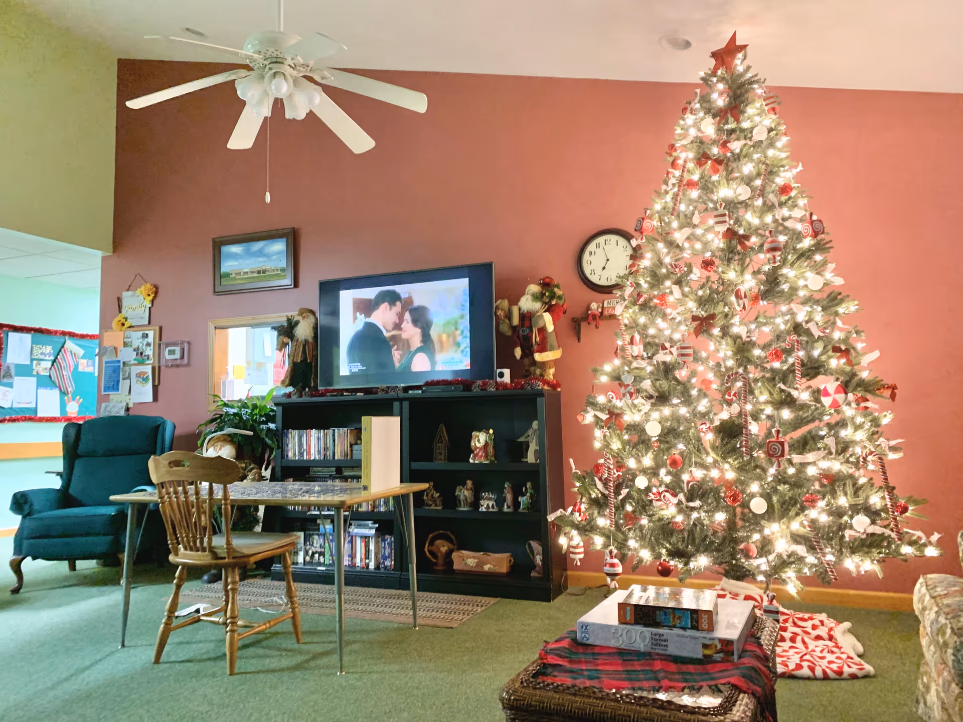 A cozy living room decorated for Christmas with a large, brightly lit Christmas tree adorned with red and white ornaments. There is a wooden chair and a small table in front of a black entertainment center holding a TV showing a romantic scene, DVDs, and holiday decorations including a Santa figure. The walls are painted red and there is a ceiling fan above. A clock and various decorations are on the wall behind the tree.