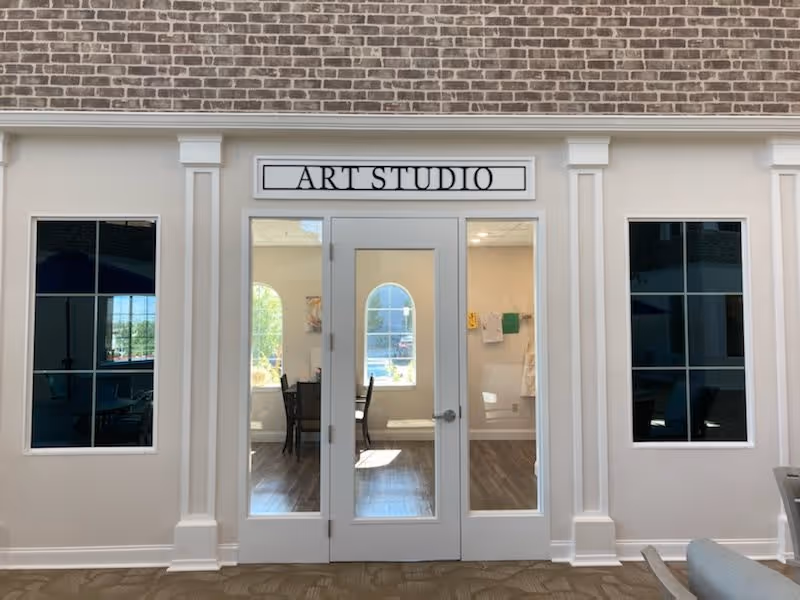Entrance to an art studio with a glass door and two large windows on either side. Inside, there are chairs and tables visible, along with a window letting in natural light. The walls are light-colored with decorative molding and a sign above the door reads 'ART STUDIO'.