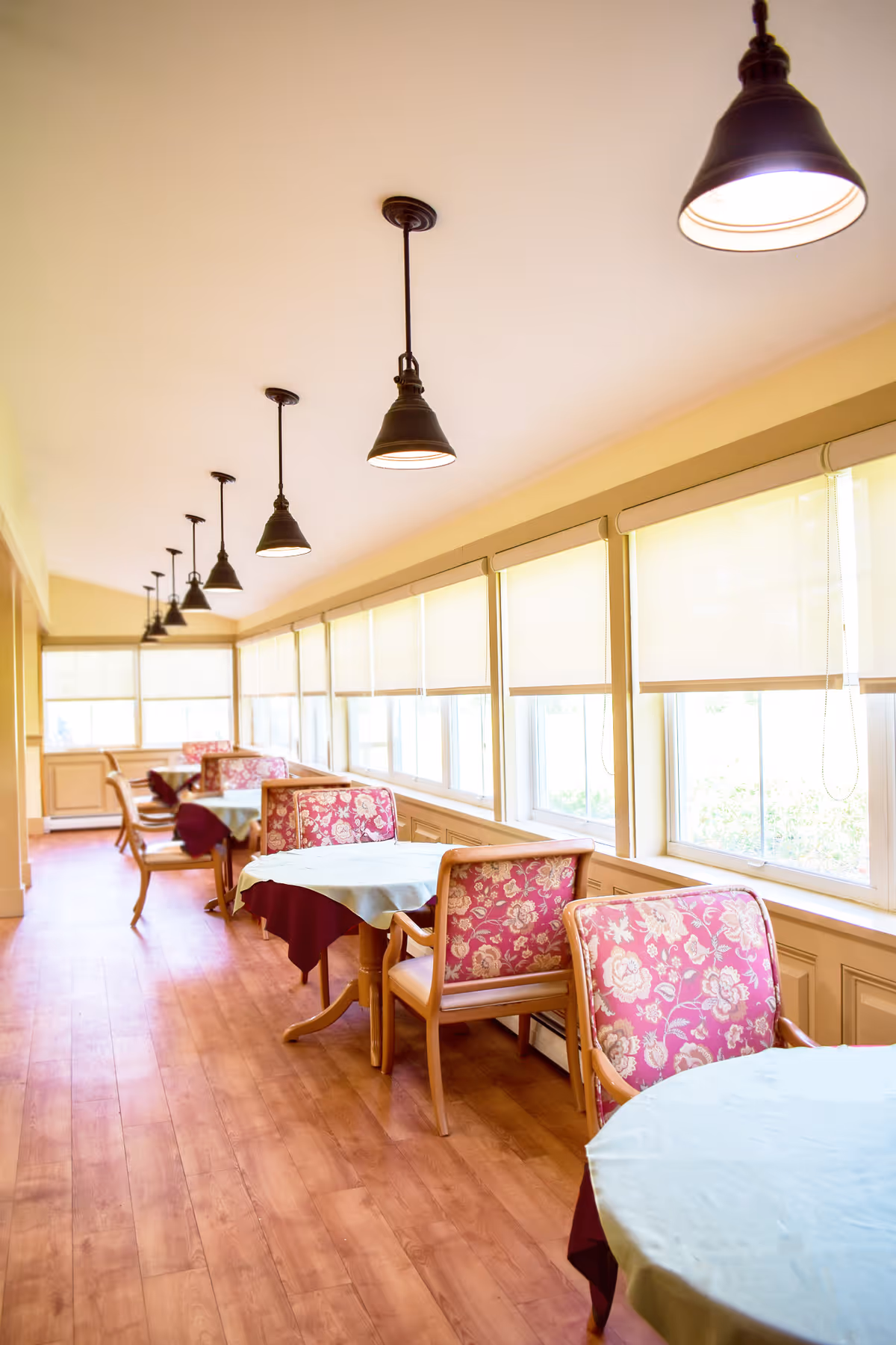 Sunlit dining room with round tables, floral-upholstered chairs, and pendant lights along a row of windows.