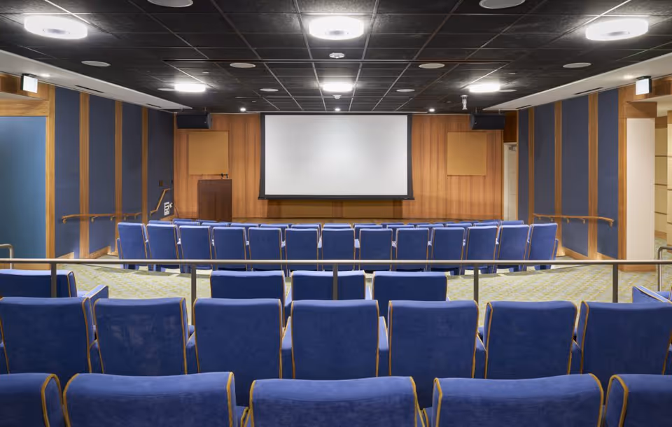 Small auditorium with rows of blue upholstered chairs facing a projection screen and a wood-paneled front wall.