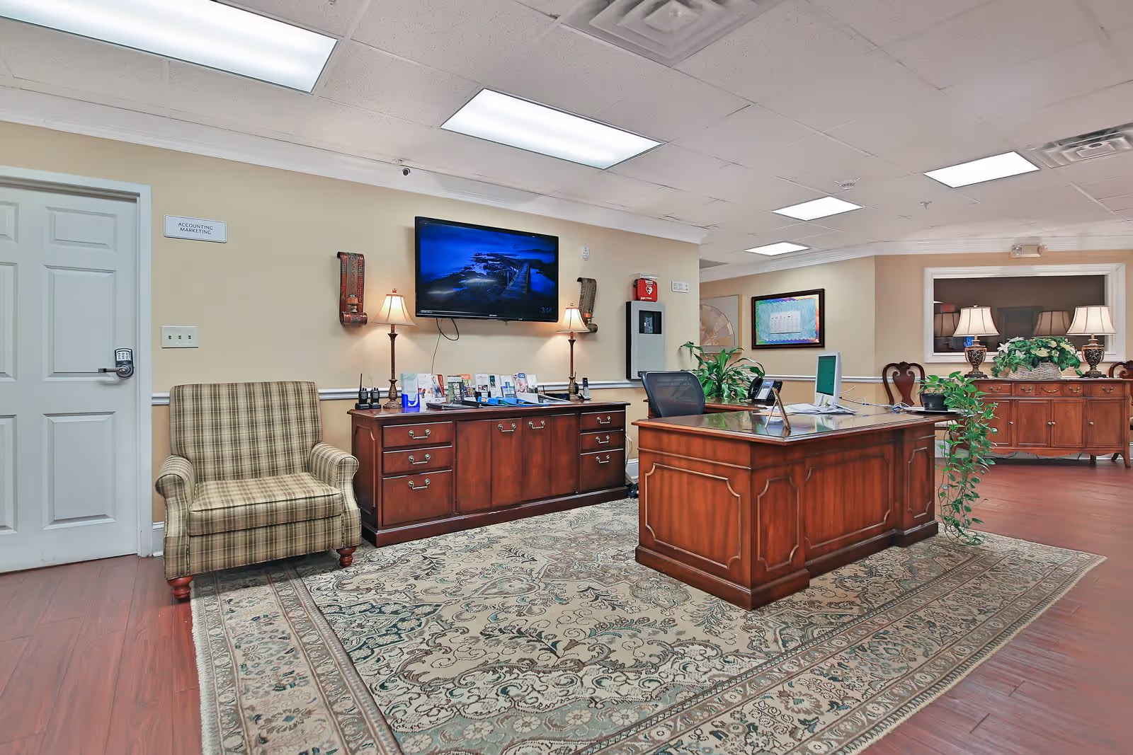 An office area inside Manchester Court Assisted Living and Memory Care featuring a wooden desk with a computer, a matching wooden cabinet with brochures and two lamps, a plaid armchair, a wall-mounted TV, plants, and a large patterned rug on a wooden floor.