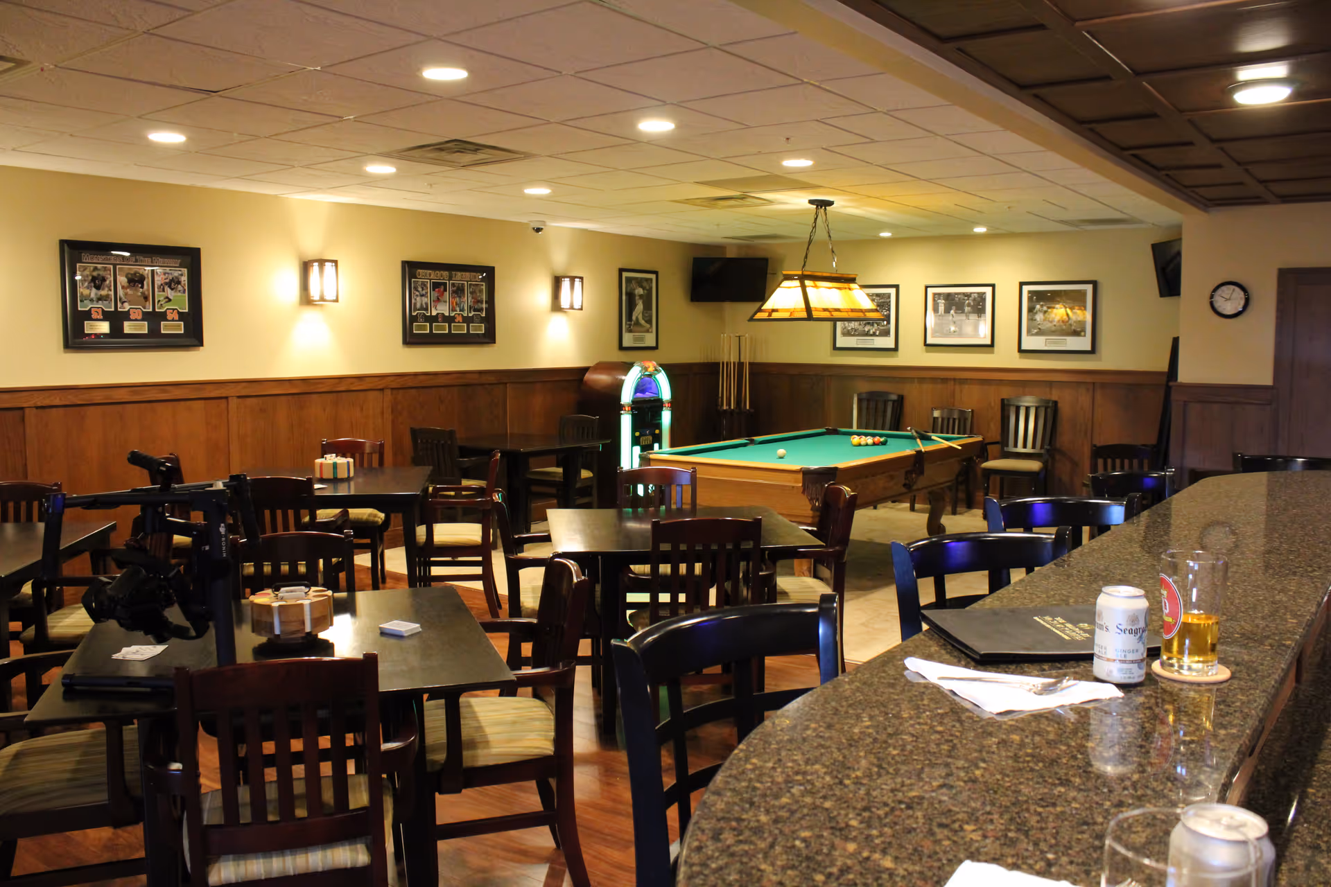 Interior view of a recreational room with multiple tables and chairs, a pool table with balls and cues, a jukebox, framed sports photos on the walls, and a granite countertop bar with drinks and napkins.