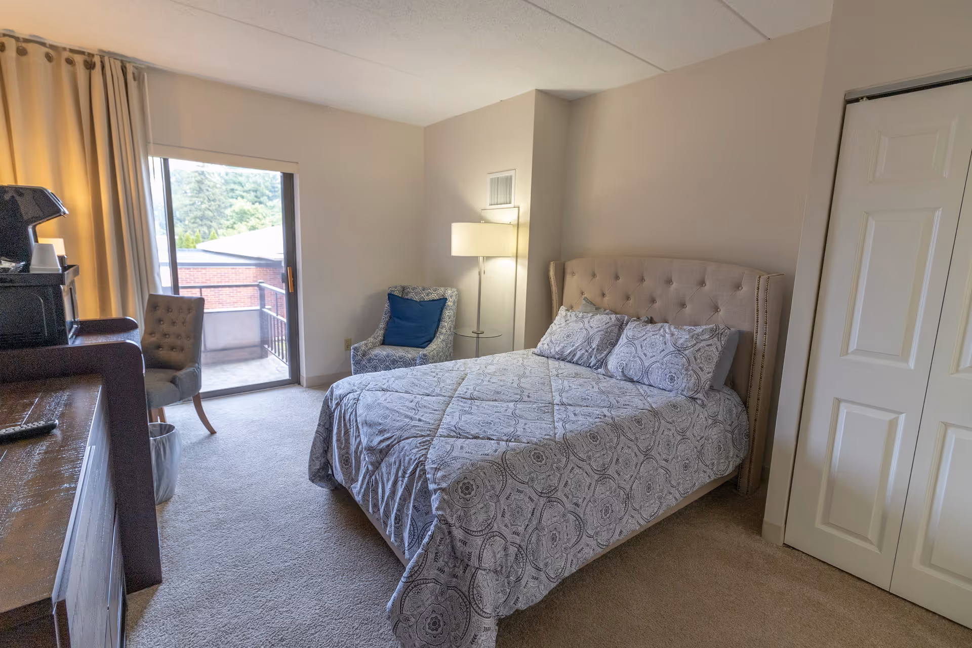 A bedroom with a bed covered in a patterned gray and white comforter, a blue patterned armchair with a blue cushion, a floor lamp, a wooden dresser with a coffee maker on top, a chair, and a sliding glass door leading to a small balcony with a view of trees and a brick building outside.