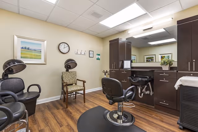 Interior view of a hair salon area in a senior living facility with salon chairs, hair drying stations, a large mirror, dark wood cabinetry, a clock on the wall, and a framed landscape painting.