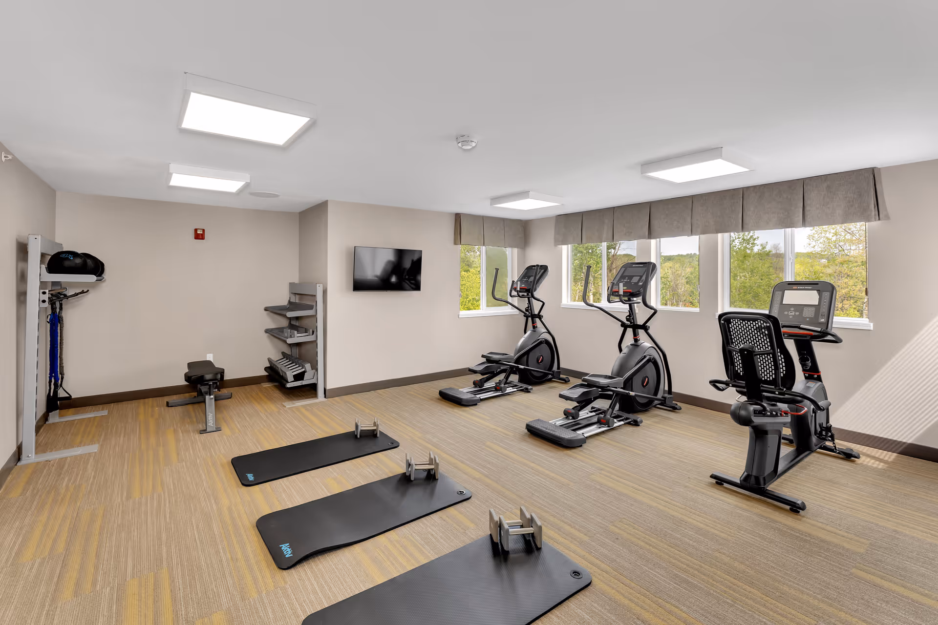 A bright fitness room with three exercise mats each paired with a set of dumbbells on a carpeted floor. The room has two elliptical machines and a recumbent bike positioned near windows with a view of greenery outside. There is a wall-mounted TV and shelves holding additional exercise equipment against a beige wall.