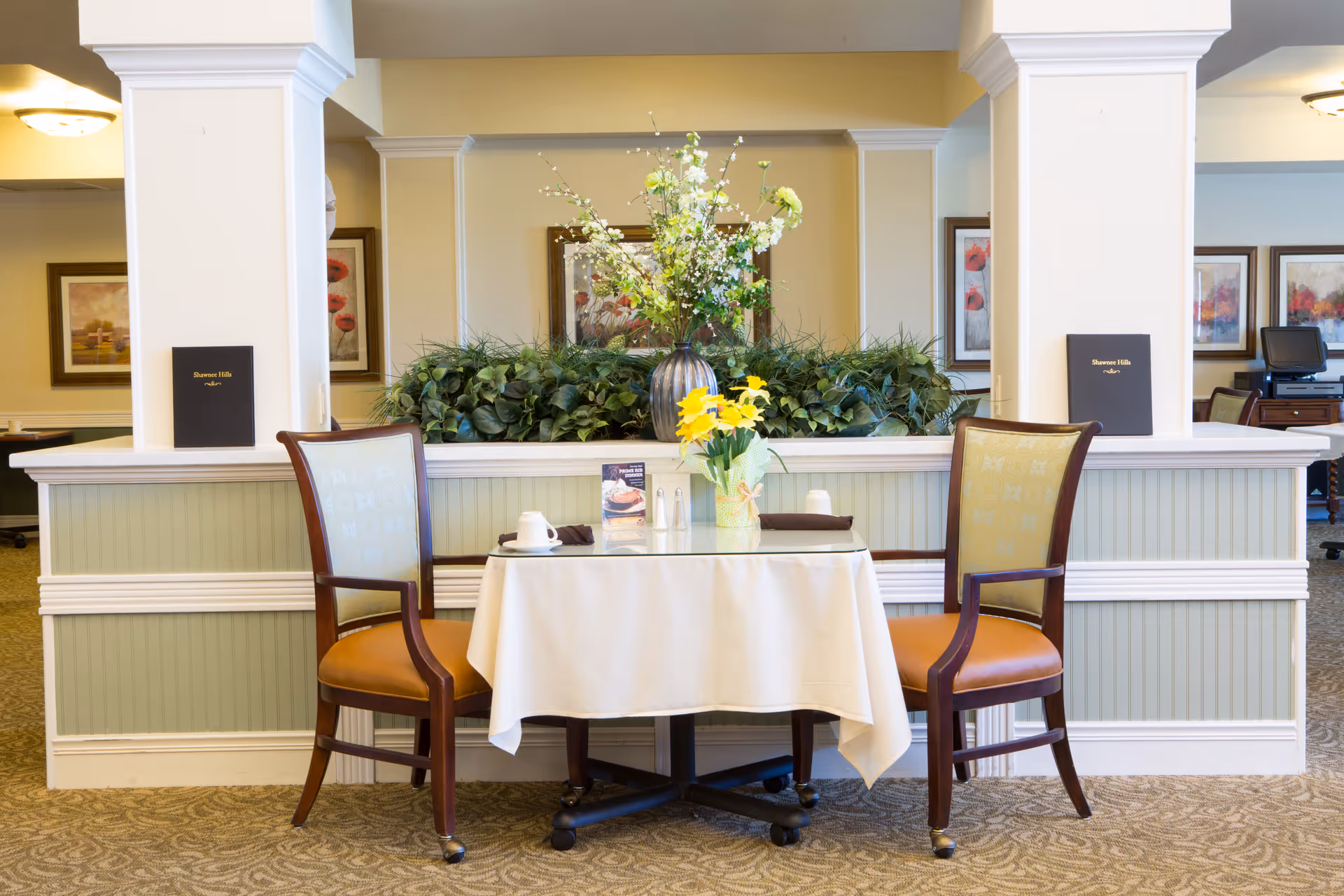 A small dining table with a white tablecloth set for two with a cup, napkins, and a small vase of yellow flowers. Two wooden chairs with cushioned seats are placed on either side of the table. Behind the table is a half wall with green plants and a large floral arrangement in a vase. The background shows framed artwork on beige walls and part of a computer workstation.