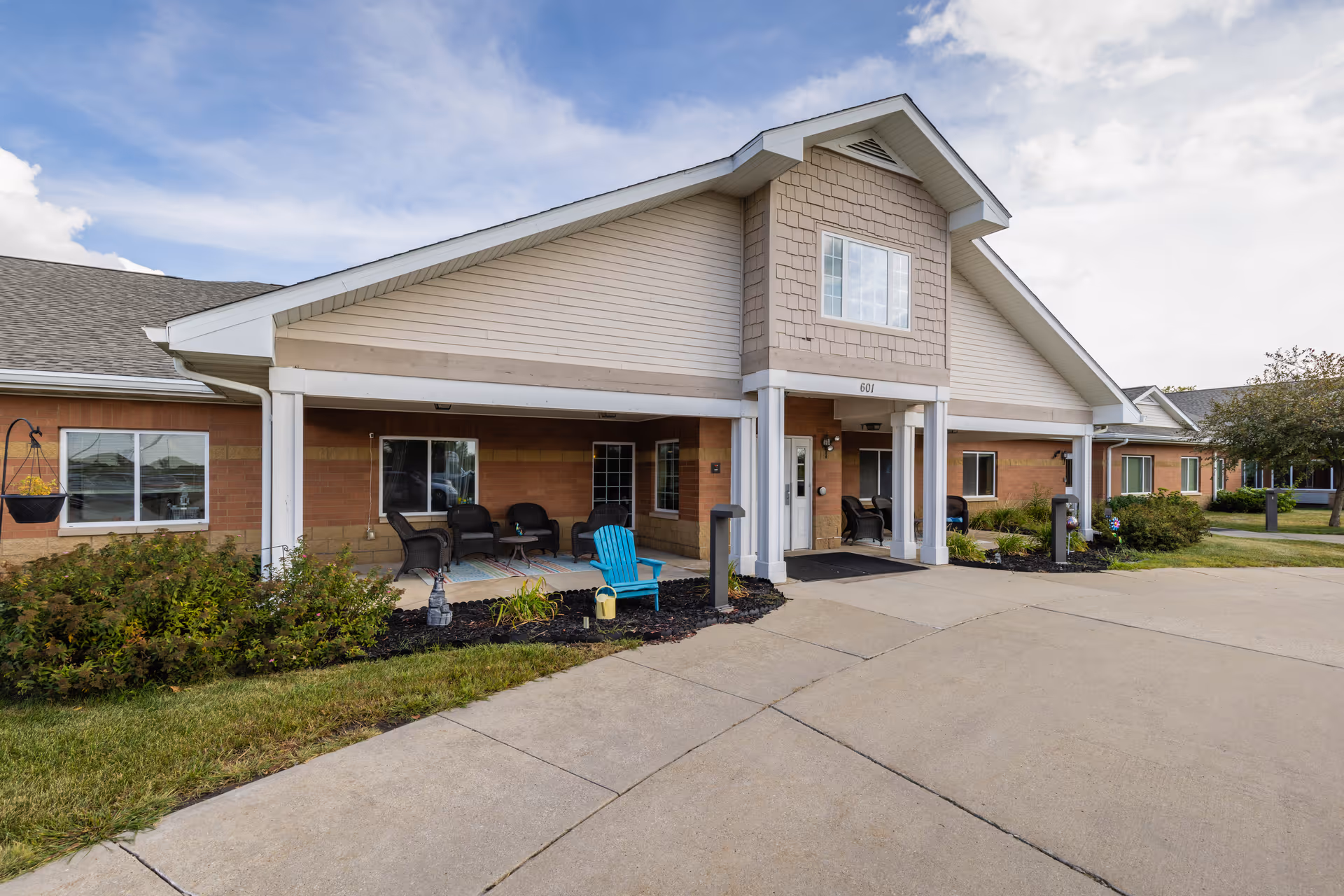 Exterior view of Courtyard Estates at Hawthorne Crossing showing a single-story building with a covered porch area furnished with outdoor chairs and a small table. The building has a combination of brick and siding with a peaked roof and several windows. There are shrubs and a lawn in front of the building along a concrete driveway.