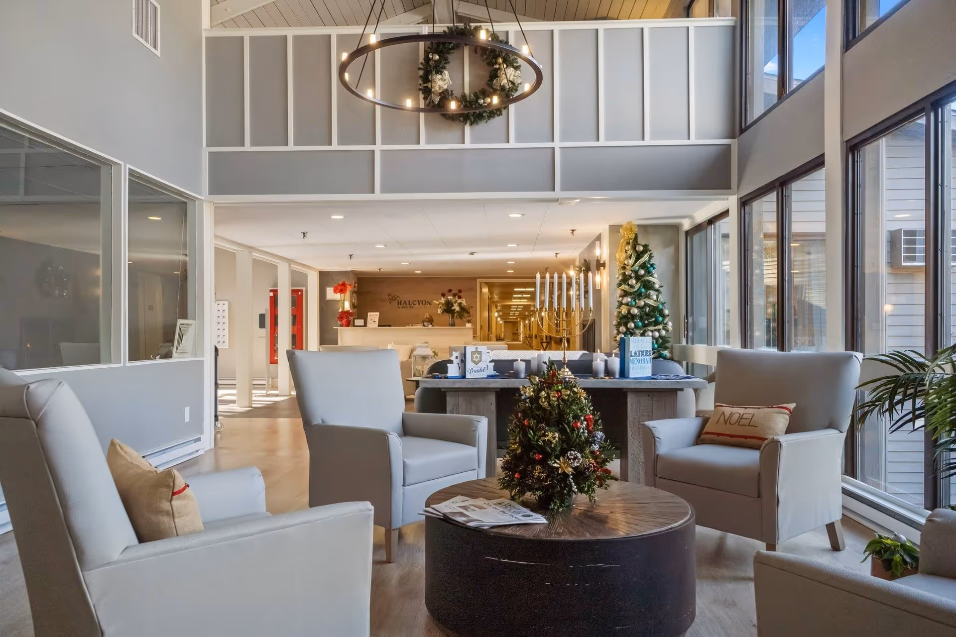 A bright and spacious senior living facility lounge area decorated for the holidays with a Christmas tree, menorah, and festive wreath. The room features comfortable armchairs arranged around a round wooden coffee table with a small decorated Christmas tree centerpiece. Large windows allow natural light to fill the space, and the reception desk is visible in the background with the Halcyon logo on the wall.