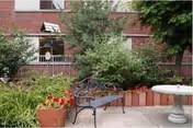 Outdoor patio area with a metal bench, potted plants with red flowers, a concrete birdbath, and greenery in front of a brick building with windows and a balcony.
